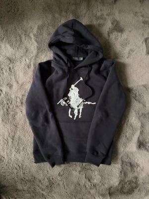 Svart hoodie från Ralph Lauren - Sny covering svart hoodie från Ralph Lauren block med stor vit polospelare och log – riktigt clean och stilren. Klassisk kängur Serv fick tier och justerbar huva med snören. Perfekt för dig som vill ha en ikonisk streetwear-look.