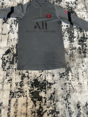 Grå PSG träningsjacka Nike - Säljer en grå träningsjacka från Nike med PSG-logga på bröstet och Accor Live Limitless-tryck. Jackan har halv dragkedja framtill, svarta detaljer på ärmarna och röd Nike-logga. Perfekt för fotbollsträning eller sport.