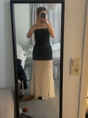 Svart och vit långklänning GinaTricot - Snygg långklänning med svart, figurnära överdel och plisserad vit/creme kjol från GinaTricot. Klänningen är axelbandslös. Aldrig använt, köpte till event, men kom aldrig till användning. Köpt för 699 kr.