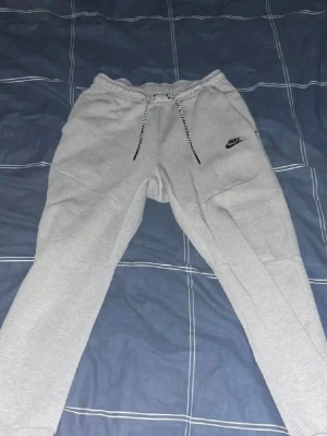 Gråa Nike mjukisbyxor med snörning - Snygga grå mjukisbyxor från Nike med svart logga på vänster ben och dragsko i midjan. Byxorna har ribbade muddar vid bensluten och praktiska fickor, varav en med dragkedja bak. Perfekta för chill eller träning.