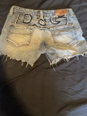 Blå jeansshorts från D&G - Säljer ett par ljusblå jeansshorts från D&G med råa, fransiga kanter och stora broderade D&G-bokstäver på bakfickorna. Klassisk femficksmodell med knappgylf och läderpatch i midjan. Perfekta för sommaren!