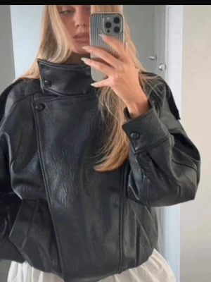 Svart skinnjacka från Zara - Cool svart skinnjacka från Zara med bred krage och asymmetrisk knäppning. Jackan har en boxy passform, två framfickor och elastisk nederkant. Perfekt för att ge din outfit en edgy touch.