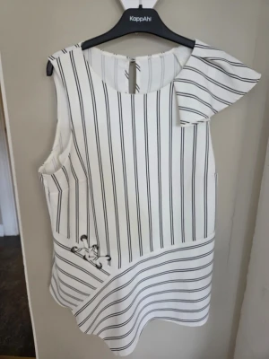 Vit randig blus med asymmetrisk volang Stl 40  - Säljer en vit blus från ZARA i Danmark stl M  med svarta vertikala ränder och en unik asymmetrisk volangdetalj vid axeln. Blusen är ärmlös och har en knapp i nacken l. Materialet känns lätt och luftigt. Snöre med öljetter fram Ena ärmen med liten volang andra ärmlös Längd bak 57 cm Längd fram där den är som längst 78 cm 97 % polyester 3 % Elastan OBS STL OCH TVÄTTLAPP BORTKLIPPT DÅ DEN KLIADE