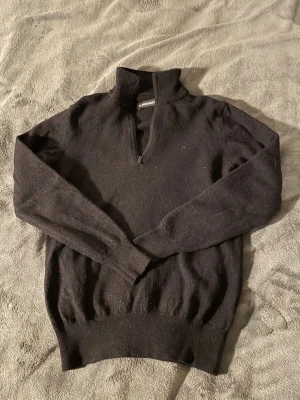 Svart stickad tröja med halv dragkedja - Svart stickad tröja från J.Lindeberg med halv dragkedja och hög krage. Tröjan har långa ärmar och ribbade muddar vid ärmslut och nederkant. Perfekt för lager på lager och enkel att matcha med olika outfits. Defekt är att märkeslappen sitter halvt av annars toppen skick. Står S men är väldigt liten i storlek👍