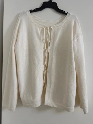 Krämvit kofta med knytband - Säljer en krämvit kofta med långa ärmar och tre dekorativa knytband framtill. Koftan har rund halsringning och ribbade muddar vid ärmslut och nederkant. Perfekt för lager på lager och enkel att styla till olika outfits.Övre knytbandet på koftan är lite löst (se bild).