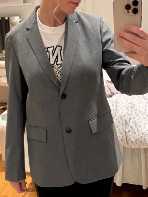 Uniqlo kavaj grå - Snygg grå kavaj från Uniqlo med två knappar framtill och klassisk krage. Rak passform och två fickor med lock. 