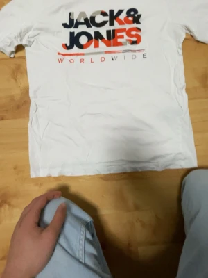 Vit soprtig Jack & Jones t-shirt - Snygg vit t-shirt från Jack & Jones med stort tryck i svart, orange och grått framtill där det står 'JACK & JONES WORLDWIDE'. Klassisk passform och rund hals. Perfekt till jeans eller shorts för en avslappnad look.