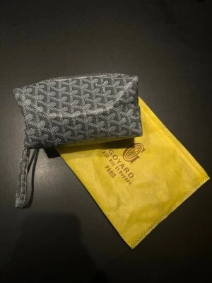 Grå Västra för necessär med mönster - Säljer en grå necessär med klassiskt vitt mönster och dragkedja. Väskan har en praktisk handledsrem och kommer med en gul dustbag med logga. Perfekt för att organisera småsaker eller smink.