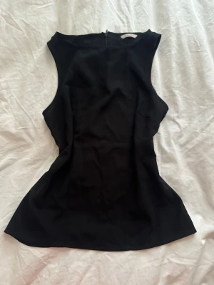 Svart ärmlös topp  - Snygg svart ärmlös topp fmed hög halsringning och figurnära passform. Toppen har dragkedja i ryggen och är tillverkad i ett slätt material som ger en stilren look. Perfekt att styla med jeans eller kjol.
