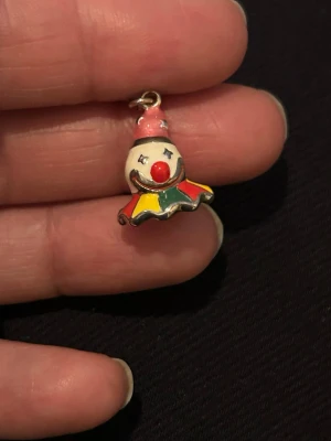 Färgglatt clownberlock i emalj - Unik berlock formad som ett clownhuvud med röd näsa, rosa hatt och krage i gult, grönt och rött. Tillverkad i silver metall med emaljerade detaljer och en liten ögla för att fästa i halsband eller armband. Perfekt för dig som gillar lekfulla accessoarer.