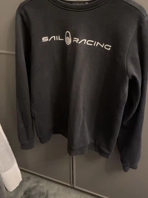  Sail Racing  tröja  - Cool svart sweatshirt från Sail Racing med vit logotyp på bröstet. Tröjan har rund halsringning och långa ärmar med ribbade muddar. Perfekt för dig som gillar sportig stil och vill ha något bekvämt och snyggt.