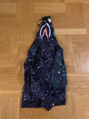 BAPE Shark hoodie med galaxmönster - Säljer en BAPE Shark hoodie med ikoniskt hajmotiv på huvan och galaxmönster över hela plagget. Huvan har broderade bokstäver i olika färger och dragkedja hela vägen upp. Perfekt för dig som vill sticka ut med streetwear-stil.
