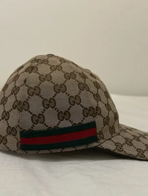 Gucci keps - Säljer en beige keps från Gucci med det klassiska GG-monogrammet över hela kepsen. På sidan finns en grön och röd rand som ger en ikonisk touch. Kepsen har böjd skärm och är tillverkad i canvasmaterial.