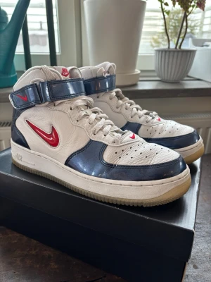 Nike Air Force 1 High blå/vit/röd stl 42,5 - Nike Air Force 1 High sneakers i vitt läder med blå detaljer och röd Swoosh. Klassisk hög modell med kardborreband runt ankeln, perforerad tåbox och robust sula. Snygg retrostil med tydliga Nike-loggor på plös och häl. De är endast använda ett par gånger och är därför i mycket bra skick. Säljes för att de är för små. Hör gärna av dig vid funderingar. Köpta på Nikes hemsida och nypris är 1600kr!