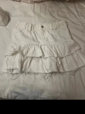 Vit volangkjol - Supersöt vit kortkjol med flera volanger och råa kanter för en trendig look. Kjolen har knapp och dragkedja framtill samt fickor. Perfekt för dig som vill ha en feminin och avslappnad stil.