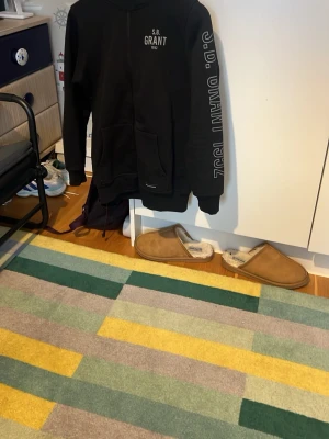 Svart hoodie från S.B. Grant - Svart hoodie från S.B. Grant med tryck på bröstet och text längs ena ärmen. Klassisk modell med huva och känguruficka framtill. Perfekt för en avslappnad och cool stil. Tillverkad i mjukt material för extra komfort.