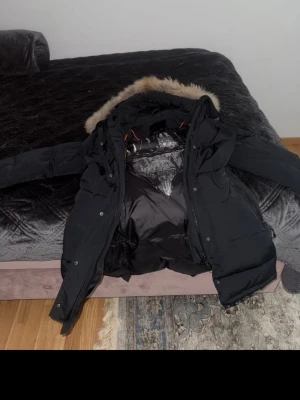 Svart parkas från Hollies med äkta päls - Snygg svart parkas från Hollies med avtagbar huva och äkta päls. Jackan har dragkedja, knappar och flera fickor. Fodrad insida med coolt bergsmotiv och orange detaljer. Perfekt för kalla dagar och riktigt varm.