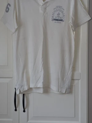 Vit Peak Performance pikétröja - Klassisk vit pikétröja från Peak Performance med tryck på bröstet och siffran 6 på ärmen. Den är i fint skick. Strl XL