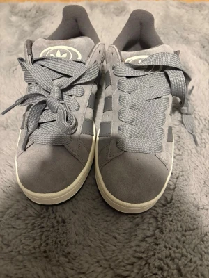 Grå Adidas Campus sneakers - Säljer ett par grå Adidas Campus sneakers i mocka med klassiska tre ränder på sidan och vit sula. Skorna har breda grå skosnören och Adidas-logga på plösen. Snygg och tidlös modell som passar till det mesta.helt nya de är inte använda / priset kan diskuteras 