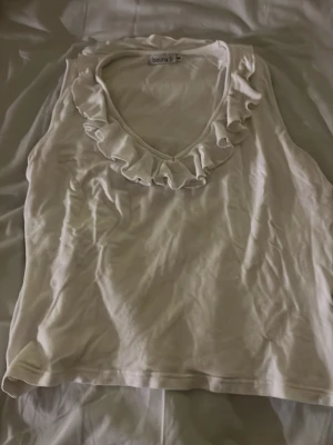 Vit linnetopp med volang från isolina  - Söt vit linnetopp från Bolina med volangdetalj runt den runda halsringningen. Toppen är ärmlös och har en enkel, stilren design som passar perfekt till sommaren. Materialet känns mjukt och lätt.