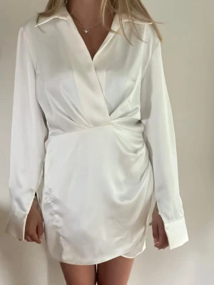 Vit satinklänning  - Stilren vit kortklänning med omlottdesign och långärmad skjortkrage. Klänningen har en lätt glansig satinlook och draperad midja som ger en snygg siluett. Perfekt för dig som gillar en chic och enkel stil. Aldrig använd 