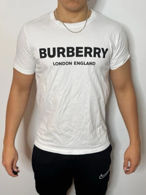 Vit Burberry t-shirt med tryck - Säljer en klassisk vit t-shirt från Burberry med svart texttryck 'BURBERRY LONDON ENGLAND' på bröstet. T-shirten har rund hals, korta ärmar och är gjord i mjuk bomull. Perfekt för dig som gillar stilrena och ikoniska plagg.