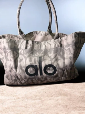 Grå tote bag från Alo - Snygg tote bag i grått med marmorerat mönster och stort svart Alo-tryck på framsidan. Väskan har dubbla handtag och är tillverkad i slitstarkt tygmaterial. Perfekt för att bära med sig allt man behöver under dagen.