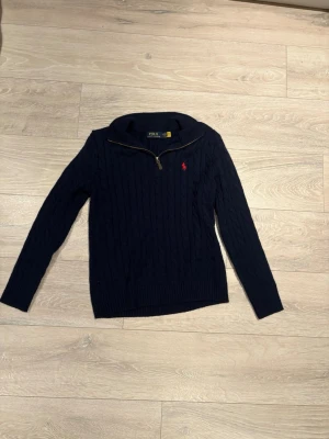 Mörkblå kabelstickad tröja Polo Ralph Lauren - Mörkblå kabelstickad tröja från Polo Ralph Lauren med halv dragkedja och röd broderad logotyp på bröstet. Tröjan har ribbade muddar och hög krage, perfekt för att hålla värmen. Materialet är mjukt och bekvämt, och designen är klassisk och stilren.
