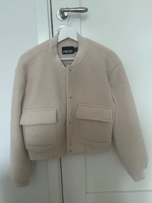 Beige jacka från PIECES - Snygg beige jacka från PIECES med boxig passform och två stora fickor framtill. Jackan har ribbade muddar vid ärmslut och nederkant samt tryckknappar framtill. Tillverkad i mjukt teddy-material som ger en mysig och trendig look.