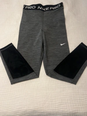 Nike Pro grå leggings med svarta detaljer - Snygga gråmelerade Nike Pro leggings med bred resår i midjan där det står 'NIKE PRO'. Svarta meshpaneler på benen och vit Nike-logga på låret. Stretchigt och mjukt material som passar perfekt till träning eller chill. Tight passform och sportig look. Aldrig använda 