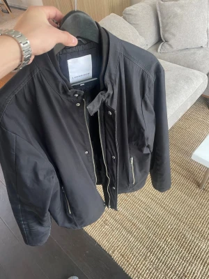 Svart bomberjacka från Lindbergh - Stilren svart bomberjacka från Lindbergh med dragkedja framtill, två sidofickor med dragkedja och ribbade muddar vid hals och ärmslut. Jackan har en klassisk bomberpassform och är tillverkad i ett slätt, tåligt material som passar perfekt till vardagen 