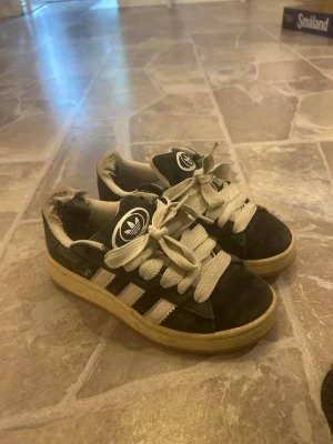 Svarta Adidas sneakers med vita ränder - Klassiska svarta Adidas sneakers med vita ränder och breda vita skosnören. Skorna har låg profil och gummisula i beige. Adidas-logga på plösen och sidan. Materialet är en mix av mocka och syntet. Perfekta för en avslappnad streetwear-look. Funkar utmärkt utöver hålen i hälen.