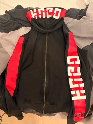 Svart hoodie med röd och vit grafik - Säljer en svart hoodie från HUGO BOSS med dragkedja framtill. Hoodien har röda paneler längs ärmarna och stor vit grafisk text på både ärmar och huva. Tillverkad i mjukt bomullsmaterial och har en klassisk huva. Perfekt för dig som gillar streetwear och tydliga detaljer.