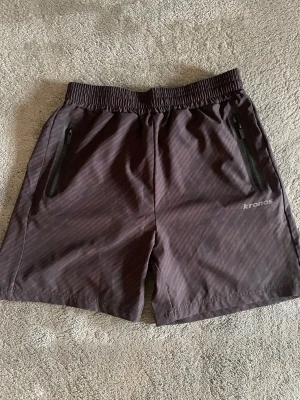 Mörkgrå träningsshorts från Kronos - Säljer ett par mörkgrå träningsshorts stretch sopass European fit från Kronos. Shortsen har diskret mönster, elastisk midja och dragkedjor på fickorna. Perfekta för sport eller gympass.