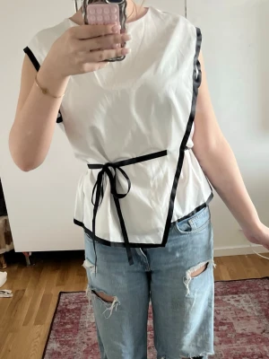 Vit omlottopp med svarta detaljer - Snygg vit omlottopp med svarta kanter och knytband i midjan. Toppen är ärmlös och har en rak, något lös passform. Materialet ser ut att vara polyester eller liknande för en lätt och luftig känsla. Perfekt att styla med jeans eller kjol. Pris kan diskuteras❤️