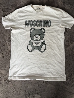 Vit Moschino t-shirt med björntryck - Säljer en vit t-shirt från Moschino med stort svart tryck av en björn och texten 'This is not a Moschino toy' på framsidan. Klassisk passform och rund hals. Perfekt för dig som gillar statement-plagg och coola prints.