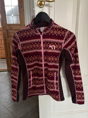 Mönstrad fleecejacka Kari Traa - Säljer en snygg fleecejacka från Kari Traa i vinrött, rosa och orange med coolt geometriskt mönster. Jackan har hel dragkedja, hög krage och två fickor fram. Perfekt för lager-på-lager och outdoor-häng. Materialet är mjuk fleece. Inga difekter alls som jag har sett