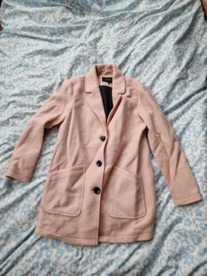 Ljusrosa kappa från Vila - Stilren ljusrosa kappa från Vila med tre svarta knappar framtill och två stora fickor. Kappan har klassisk krage och är helfodrad med svart tyg på insidan. Perfekt för dig som vill ha en enkel men snygg look till hösten eller våren.