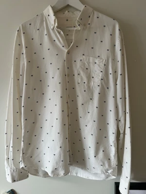 Vit skjorta från Wood Wood - Vit skjorta från Wood Wood med små svarta W-broderier över hela plagget. Mycket bra skick, stilren och fin nu till vår och sommar!
