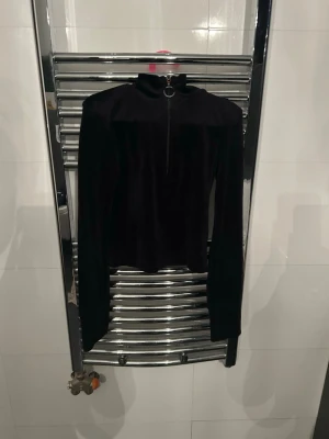 Svart sammetstopp med dragkedja - Snygg svart långärmad topp i sammet med hög krage och dragkedja framtill med ringdetalj. Perfekt att styla med jeans eller kjol för en cool look.
