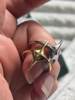 Unik tvåfärgad stjärna ring  - Säljer en cool och modern stål ring i både guld- och silverfärg. Ringen har en futuristisk, spetsig design med två sammanlänkade delar som ger en edgy look. Perfekt för dig som gillar att sticka ut med dina accessoarer.