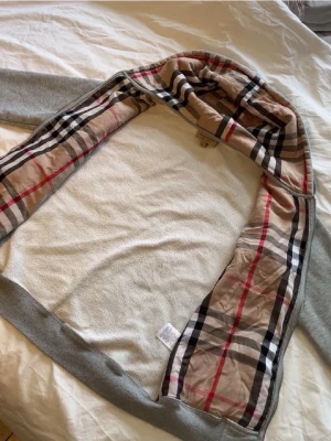 Grå hoodie med Burberry-mönster - Snygg grå hoodie från Burberry med det klassiska rutmönstret på insidan av huvan och längs öppningen. Utsidan är enkel och stilren medan insidan har beige, svart och röd design. Perfekt för dig som vill ha en hoodie med exklusiv känsla och ikonisk look.
