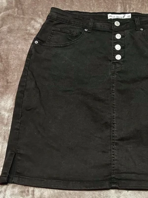 Svart jeanskjol från Blonde Story - Snygg svart jeanskjol från Blonde Story med knappgylf framtill och klassiska fickor. Kjolen har en rak passform, bälteshällor och en liten slits i sidan för extra stilpoäng. Perfekt att matcha med en crop top eller hoodie.