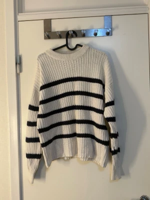 Vit stickad tröja med svarta ränder - Säljer en ribbstickad vit tröja med breda svarta horisontella ränder. Tröjan har rund halsringning, långa ärmar och avslappnad passform. Perfekt för dig som gillar klassisk och enkel stil.
