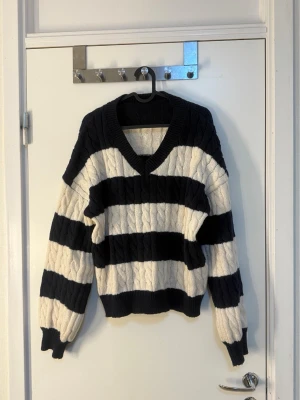 Svartvit kabelstickad tröja - Säljer en kabelstickad tröja med breda svarta och vita ränder. Tröjan har v-ringning, långa ärmar och ribbstickade muddar. Perfekt för dig som gillar klassiska mönster och en avslappnad look.