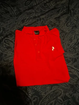 Röd Peak Performance pikétröja S - Klassisk röd pikétröja från Peak Performance med liten vit logga på bröstet och knappar framtill. Tröjan har krage och är tillverkad i ett mjukt material som passar perfekt till en avslappnad stil.