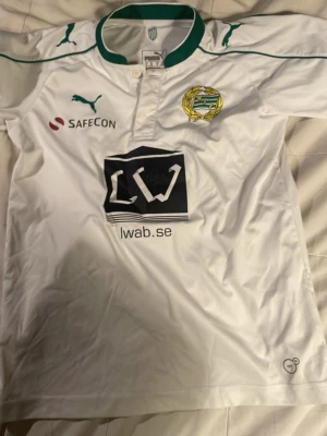 Vit Hammarby matchtröja från Puma retro - Säljer en vit Hammarby fotbollströja från Puma med gröna detaljer och klubbmärke broderat på bröstet. Tröjan har sponsorloggor och en klassisk krage i grönt. Tillverkad i lätt och funktionellt material, perfekt för träning eller match. Från 148 Cm till 156 cm