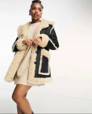 Svart fårskinnsrock med fuskpäls - Snygg svart rock med krämvit fuskpäls längs kanter, krage och ärmslut. Rocken har stora fickor framtill och en mysig, oversized passform. Perfekt för kalla dagar och ger en cool vintagekänsla.