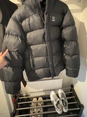 Svart pufferjacka från Haglöfs - Snygg svart pufferjacka från Haglöfs med stor huva och breda, vadderade paneler. Jackan har dragkedja framtill och diskret logga på bröstet. Perfekt för kalla dagar när du vill hålla värmen och ändå se stilren ut.
