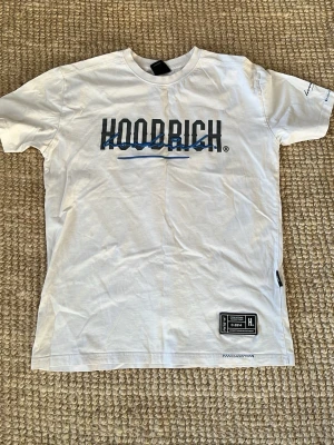 Vit HOODRICH t-shirt med blå detalj - Säljer en vit t-shirt från HOODRICH med svart logga och blå 3D-broderad text över bröstet. T-shirten har korta ärmar, rund hals och en patch med logga nertill. Perfekt för dig som gillar streetwear och snygga detaljer.